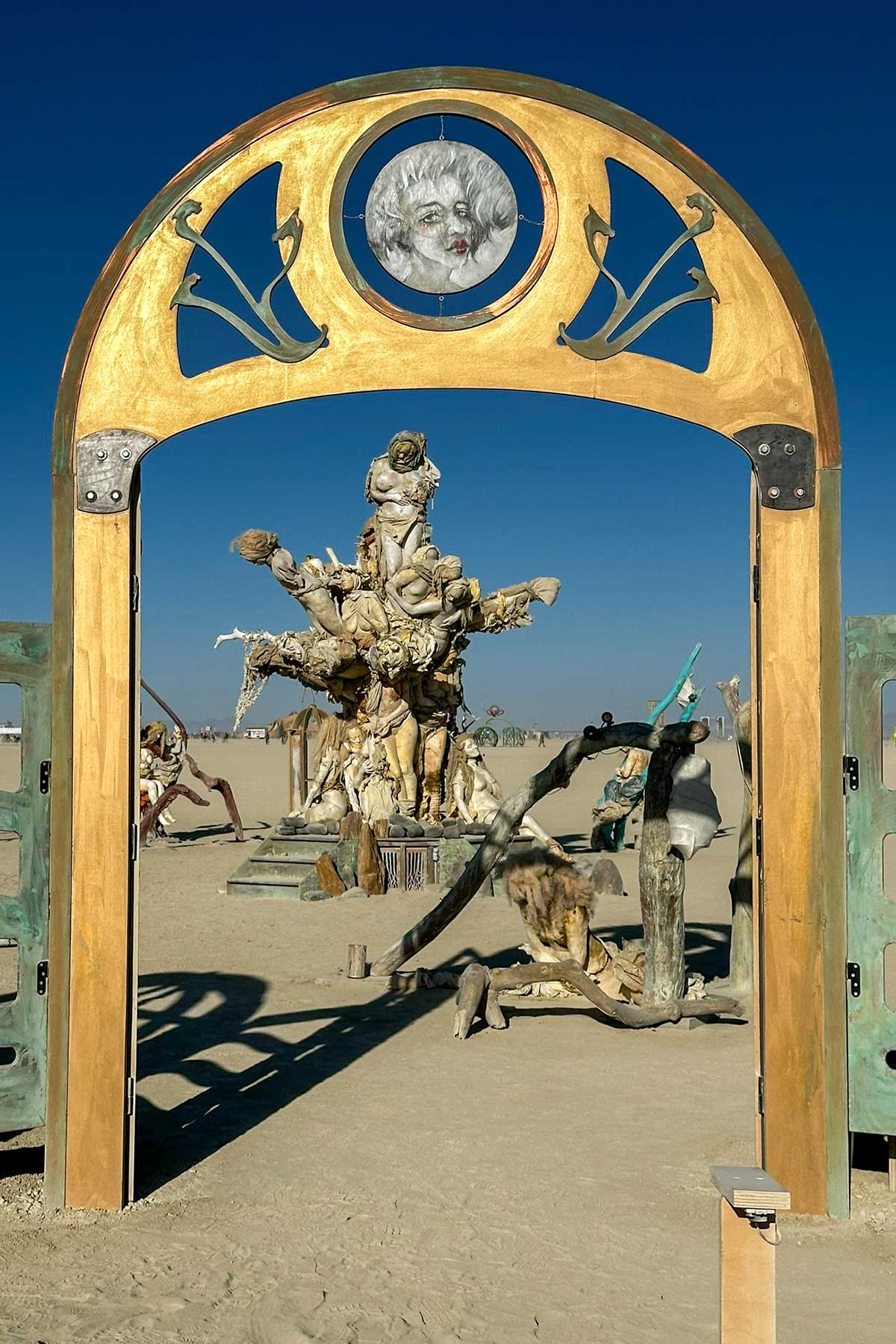 Golden arched gateway topped by a sculpted face medallion, framing Womantree on the playa under a blue sky