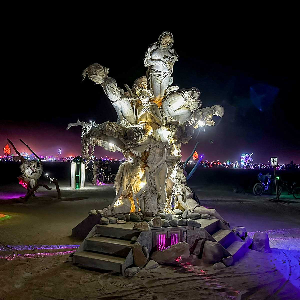 Womantree lit from within at night, its steps glowing at the base and Black Rock City lights behind