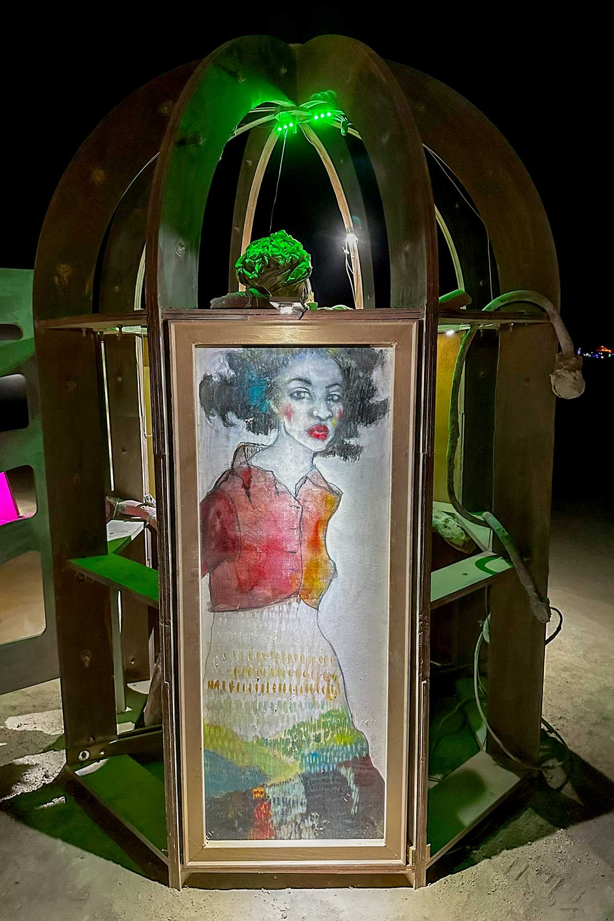 Domed kiosk in green light at night, its door showing a painted portrait of a woman in a red top and colorful skirt