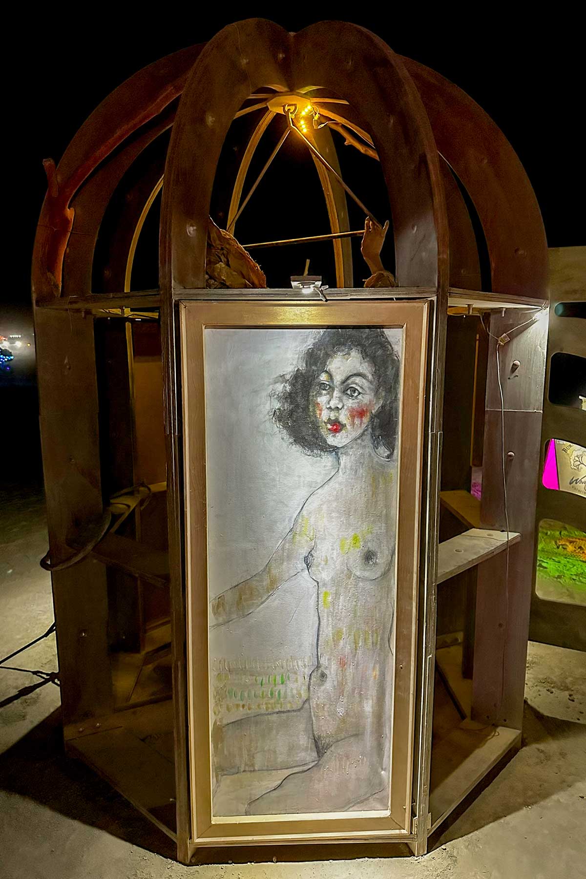 Domed kiosk in warm light at night, its door showing a painted portrait of a nude woman with curly dark hair