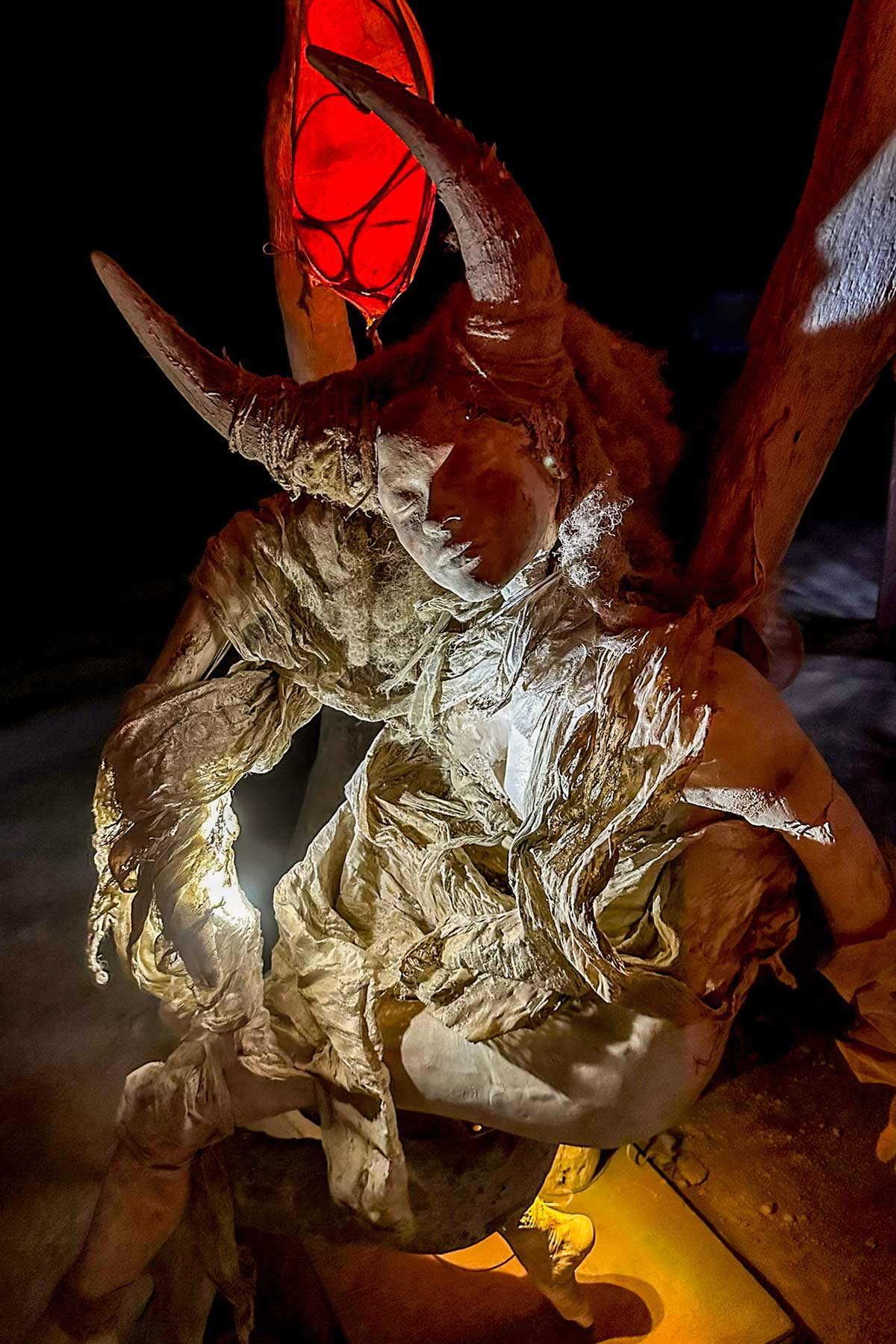 Horned cast figure in tattered fabric on a swing, lit from below with a red lantern overhead