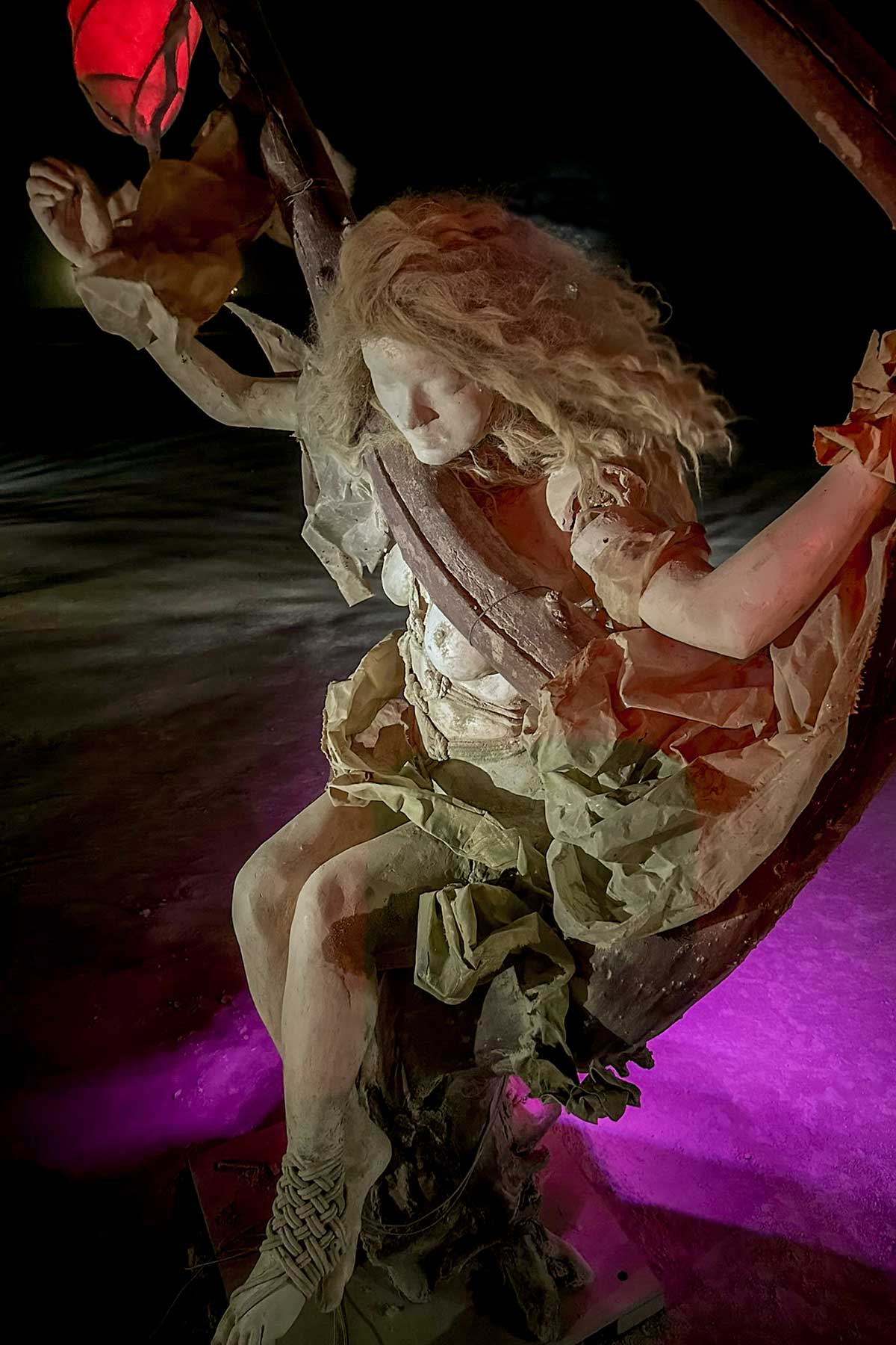 Cast figure of a woman with blond hair on a swing at night, lit in purple and pink