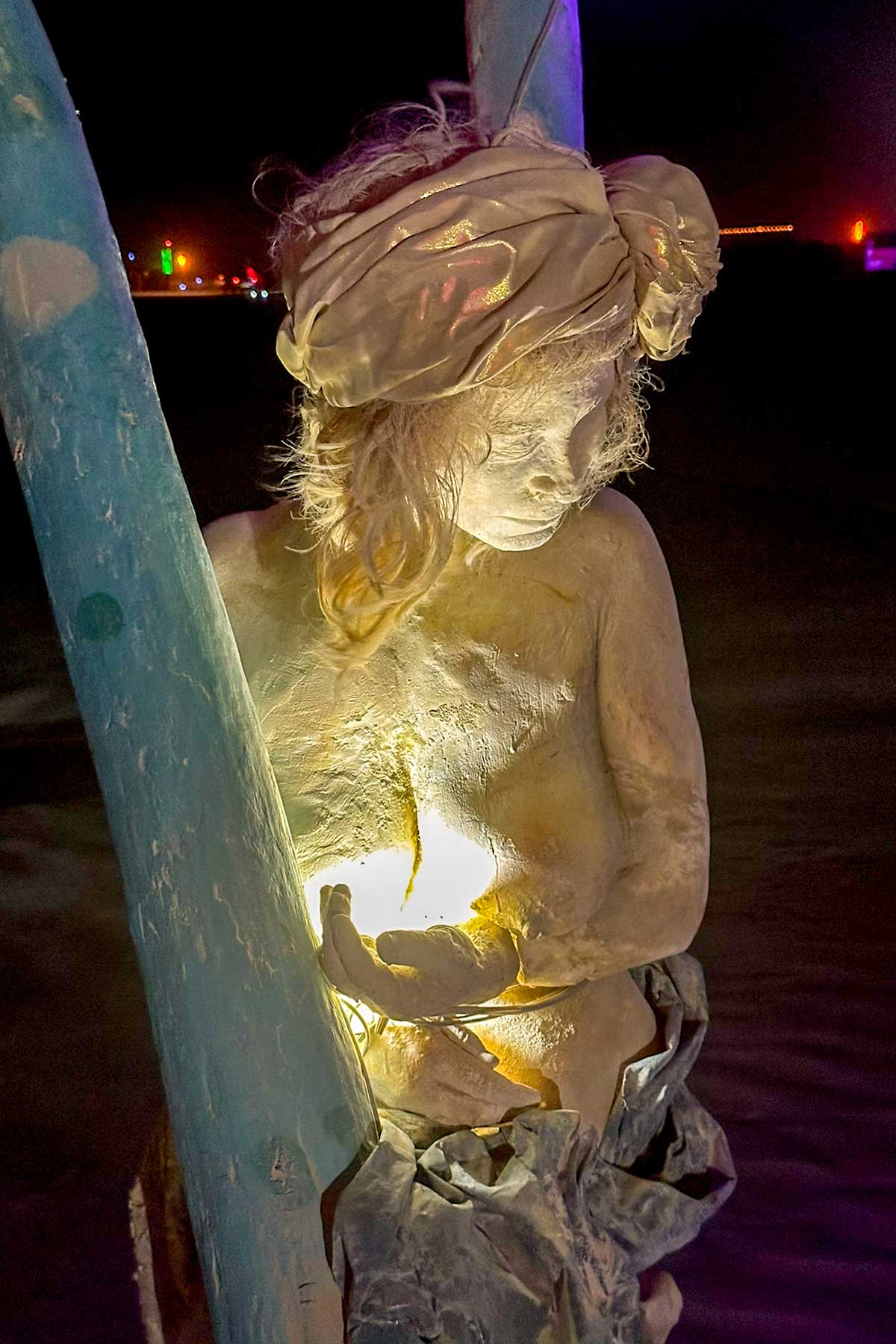 Cast figure of a woman in a head wrap on a teal swing at night, light glowing from her cupped hands