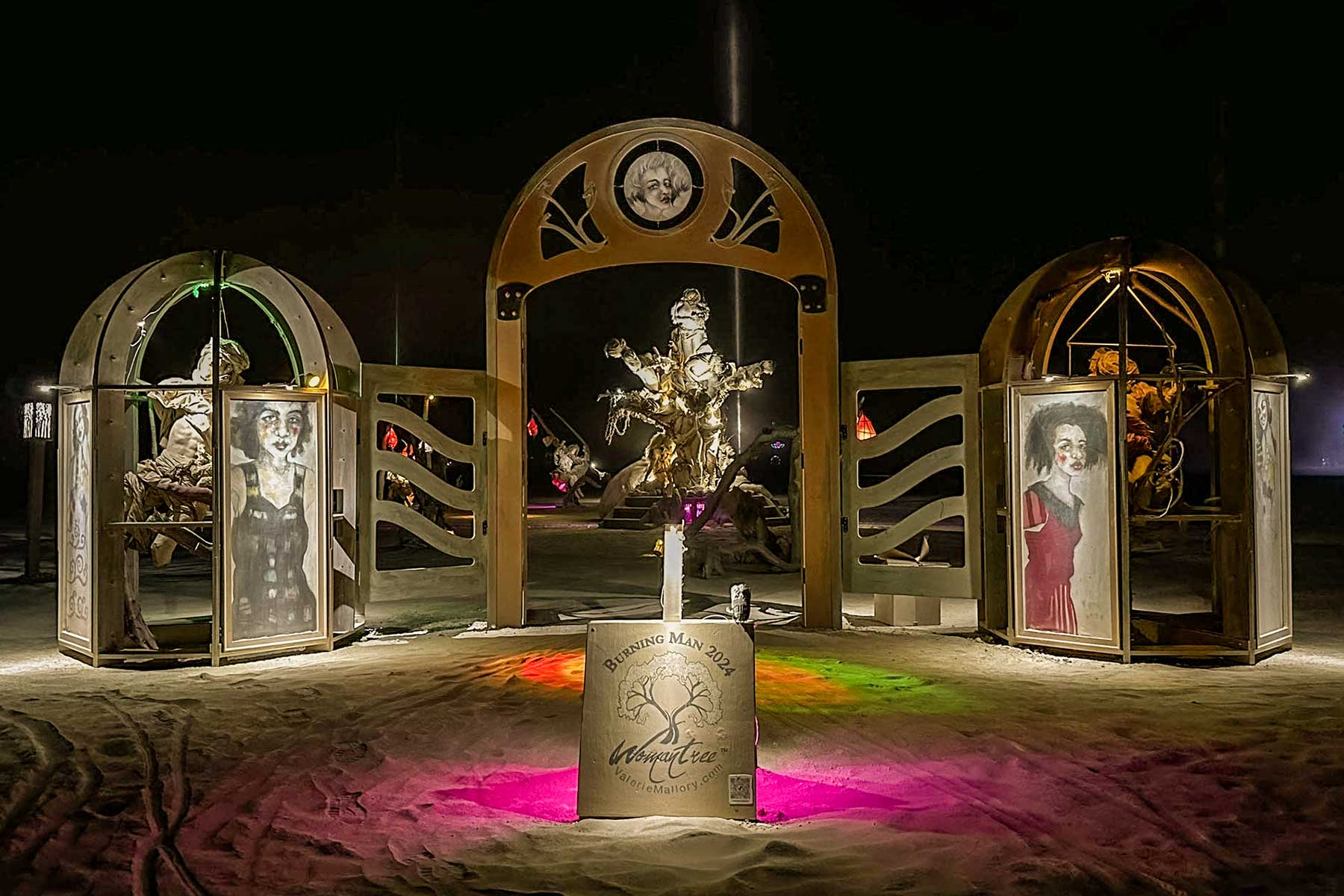 Womantree entrance at night with the gateway, two portrait kiosks and a sign, pink and green light on the sand